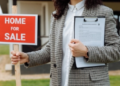 A person holding a clipboard and a home for sale sign.