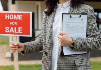 A person holding a clipboard and a home for sale sign.