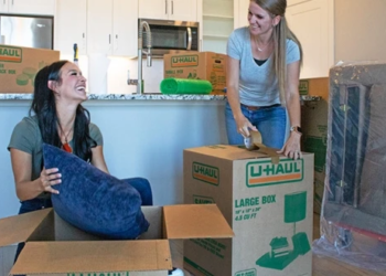 Two women laugh together as they work on packing items from their living room into U-Haul moving boxes to prepare for moving day